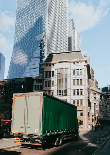 A large green and beige box truck parked on a city street near tall modern skyscrapers and historic buildings, with a clear blue sky overhead. The truck is positioned on the road, with the rear doors 